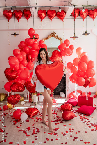 Young woman smiling while standing in room decorated for celebration Valentines Day. Girl holds large red heart shaped balloon. Rose petals scattered on floor along with pillows gift boxes. Mock up