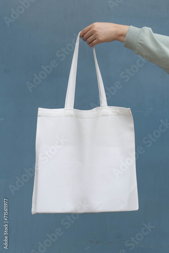 Blank white tote bag canvas fabric with handle mock up design. Close up of woman hand holding eco or reusable shopping bag on blue metal wall. No plastic bag and ecology concept. Vertical.