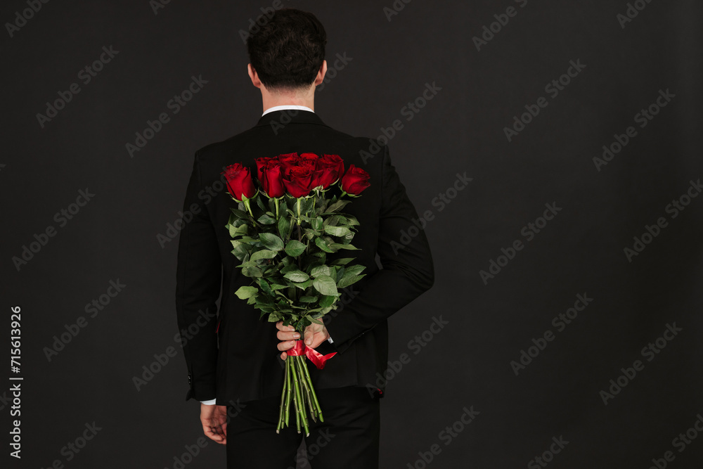 Handsome yound man in suit is standing with red roses behind the back on grey background. Valentines day. Handsome yound man in suit is standing with red roses behind the back on grey background. Valentines day.