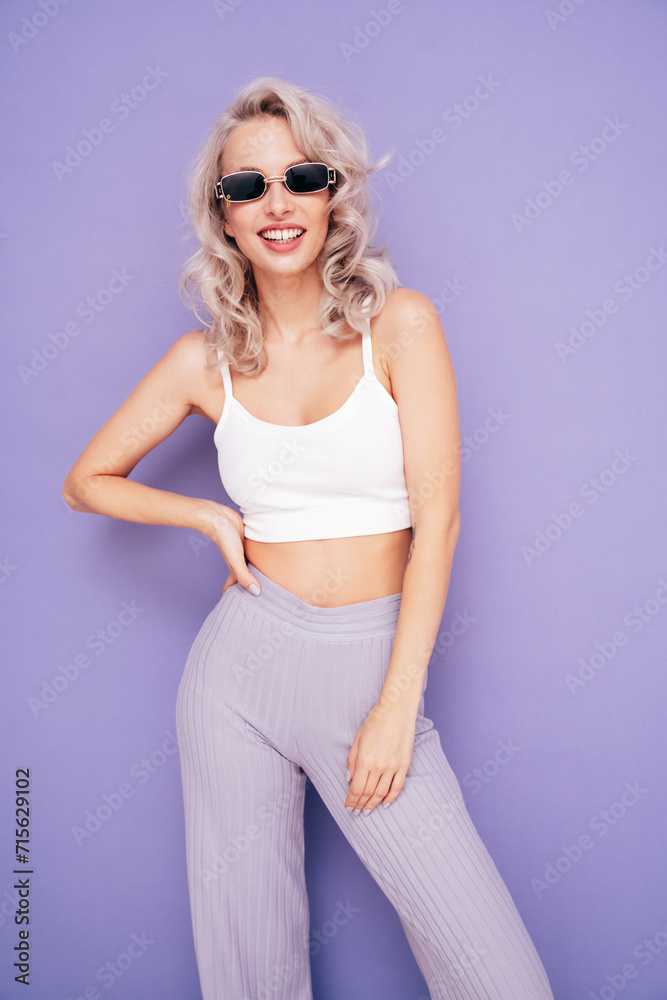 Young beautiful smiling female in trendy summer white top and pants clothes. Carefree blond woman posing near violet wall in studio. Positive model having fun. Cheerful and happy Young beautiful smiling female in trendy summer white top and pants clothes. Carefree blond woman posing near violet wall in studio. Positive model having fun. Cheerful and happy