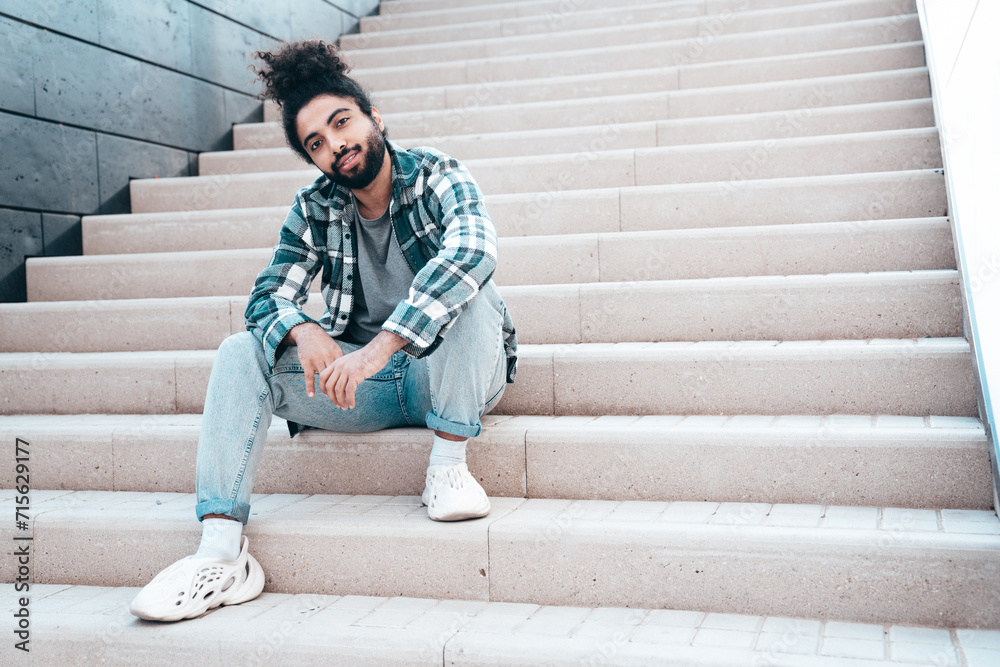 Handsome smiling hipster model. Unshaven Arabian man dressed in summer casual clothes, jeans and shirt. Fashion male with long curly hairstyle posing in the street. Sitting at the stairs