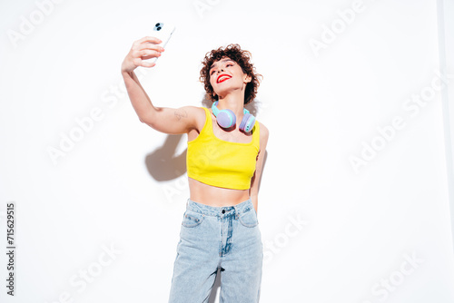 Young beautiful smiling female. Carefree woman posing near white wall in studio with curly hairstyle. Positive model having fun. Listens music, headphones on neck. Takes selfie photos