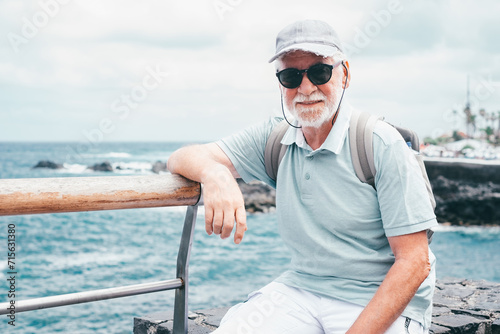 Portrait of senior traveler man with cap and sunglasses sitting at sea, elderly white haired bearded male carrying backpack enjoying vacation © luciano