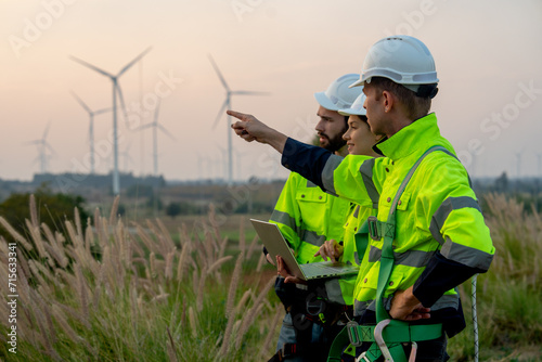 Side view and close up of technician workers man and woman work using laptop and one also point forward and stay in front of windmill or wind turbine with evening light.