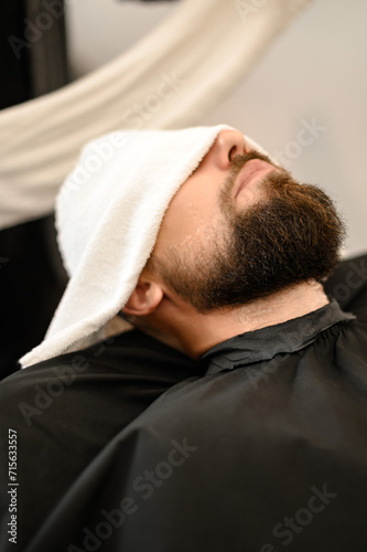 A client is lying on a chair in a barbershop with his beard wrapped in a towel. A towel covers the man eyes.