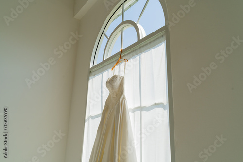 Elegant wedding dress hanging against morning light, hanging by a window in preparation of the ceremony. Symbolizing the start of a romantic wedding day