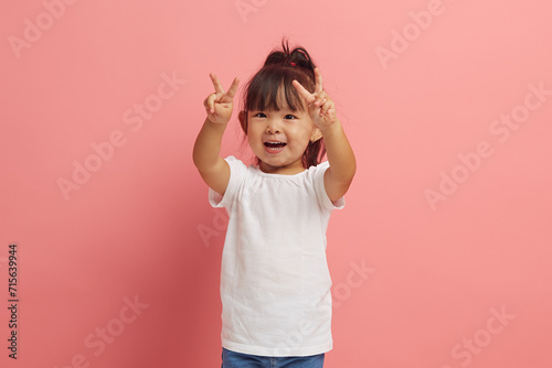 little asian etnicity funny girl showing V gesture with two hands standing over pink isolated background.  photo