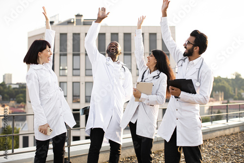 Multinational conference of doctors outdoors on rooftop of modern clinic. Group of medics celebrating success by raising their hands in team sign during break outside hospital.