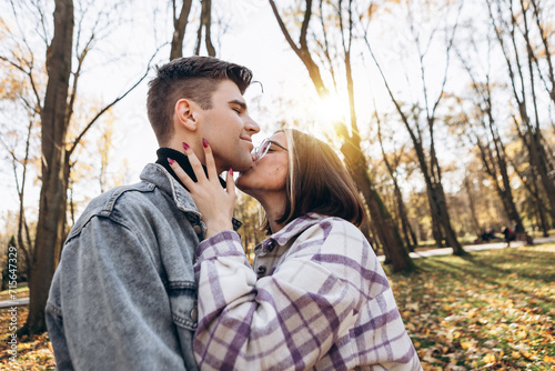 Heterosexual caucasian young loving couple walking outside in the city park in sunny weather, hugging smiling kissing laughing spending time together. Autumn, fall season, orange yellow red leaves 