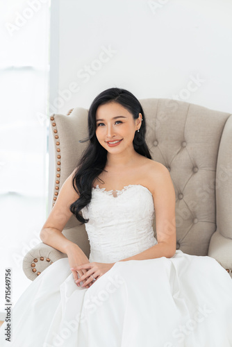 A beautiful, smiling Southeast Asian bride in white wedding gown sitting on sofa. Wedding beauty portrait in asia concept