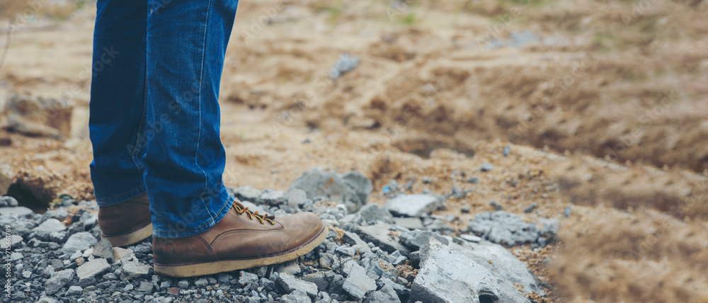 Legs of Lonely man wearing jeans and leather boots walking along the path strewn with rocks. Travel Concept-banner image.