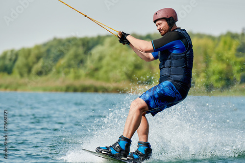a handsome man on a wakeboard in a helmet rides through the water with splashes and holds on to the winch