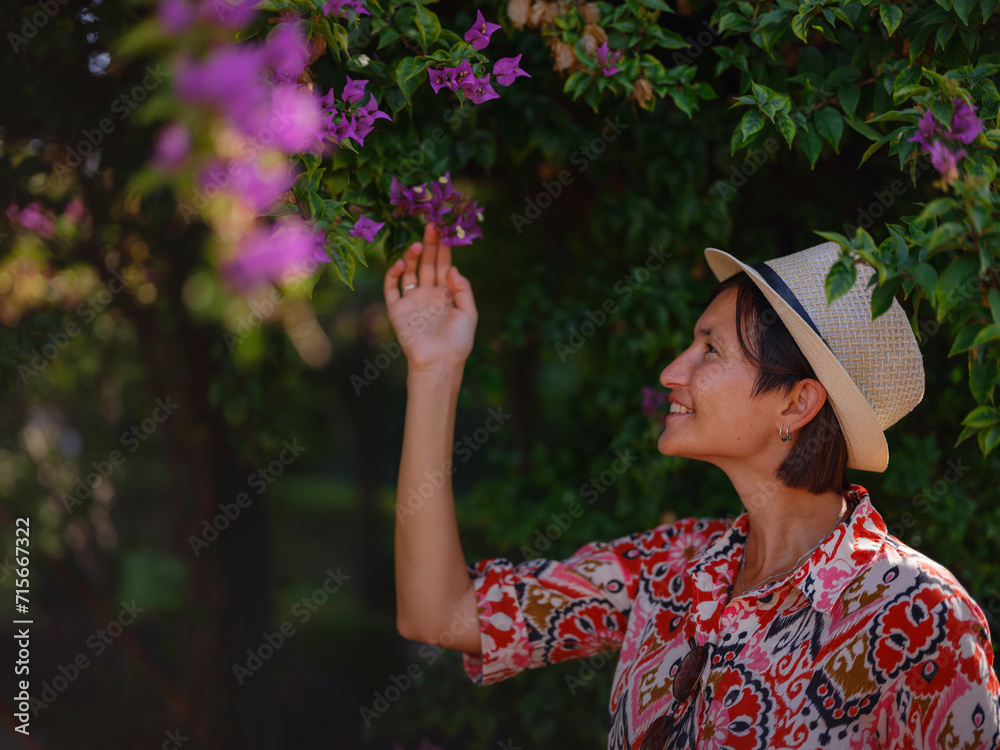 young asian woman stands in sunny green park, admiring blooming bougainvillea bush. The flower's bright colors are beautiful contrast to park's lush greenery.