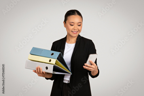 Organized Asian businesswoman carrying colorful folders and using a smartphone