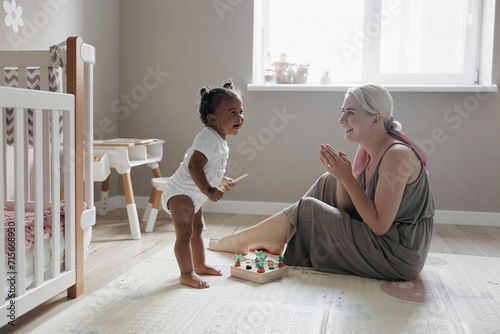 Joyful caucasian mother sits in nursery and plays with her laughing mixed race daughter.