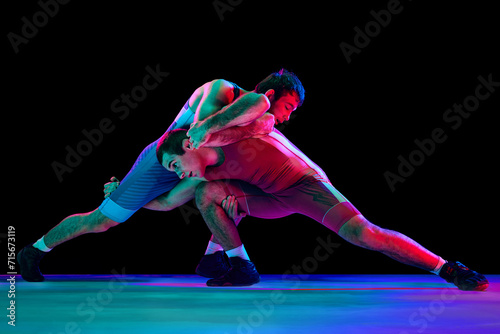 Two young athlete man, skilled wrestlers in red and blue uniform wrestling in motion against black background in mixed neon lights. Concept of motion, action, combat sports, strength, power, movement.