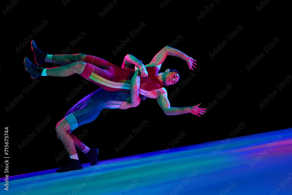 Wrestler in blue uniform lifts opponent in red, both in dynamic bridge position against black background in mixed neon lights. Mixed martial art. Concept of motion, action, combat sports, movement. ad
