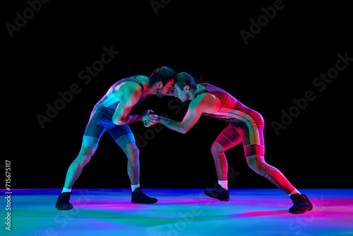 Young athlete man, wrestlers in blue and red uniform hand wrestling in neutral position on their feet in neon lights against black background. Concept of fair wrestling, championship, win competition. photo