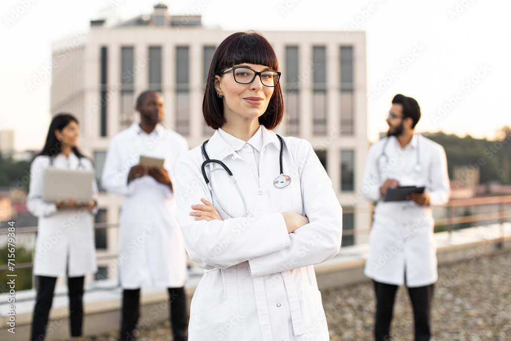 Portrait of confident Caucasian doctor in white lab coat posing on camera with crossed hands and smiling. International colleagues of mixed nationality standing behind and operating modern gadgets. Portrait of confident Caucasian doctor in white lab coat posing on camera with crossed hands and smiling. International colleagues of mixed nationality standing behind and operating modern gadgets.