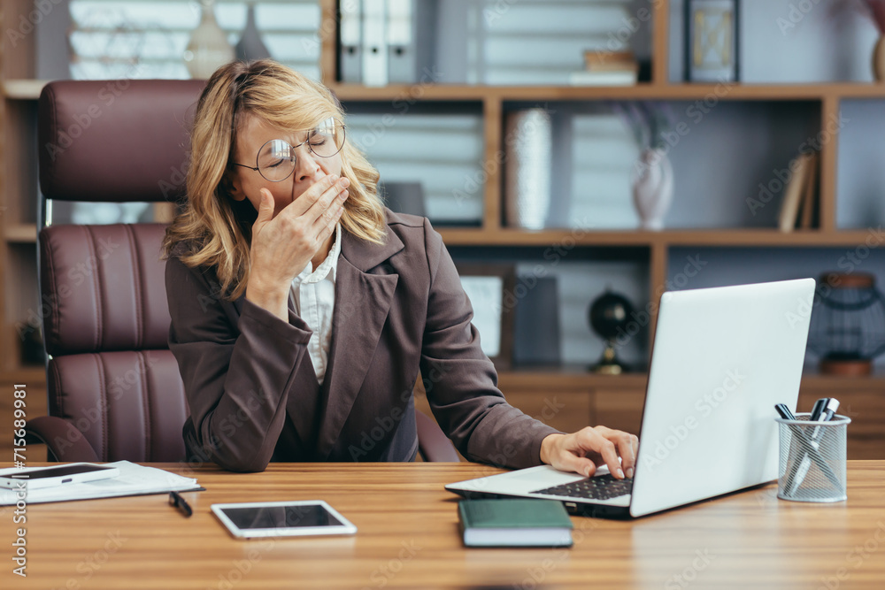 Exhausted senior blonde businesswoman in glasses yawning while working on her laptop in a well-furnished modern home office. Exhausted senior blonde businesswoman in glasses yawning while working on her laptop in a well-furnished modern home office.