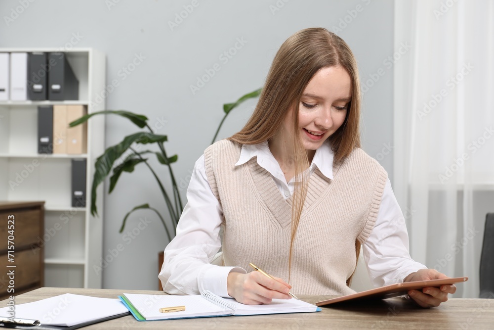 Woman taking notes while using tablet at wooden table in office, space for text Woman taking notes while using tablet at wooden table in office, space for text