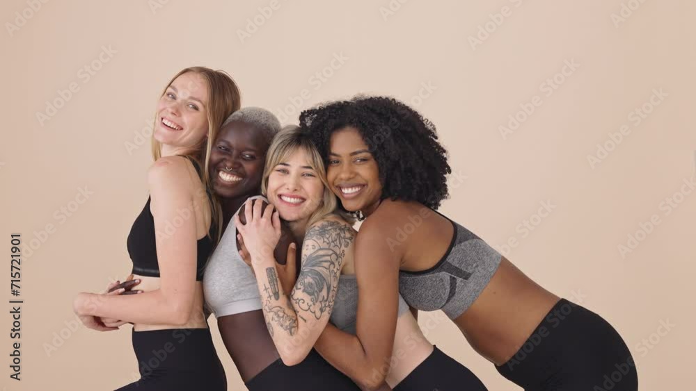Happy fit sporty diverse young women wearing sportswear hugging standing at beige background. Multiracial girls friends advertise sport fitness gym trainings together embracing posing for portrait. Happy fit sporty diverse young women wearing sportswear hugging standing at beige background. Multiracial girls friends advertise sport fitness gym trainings together embracing posing for portrait.