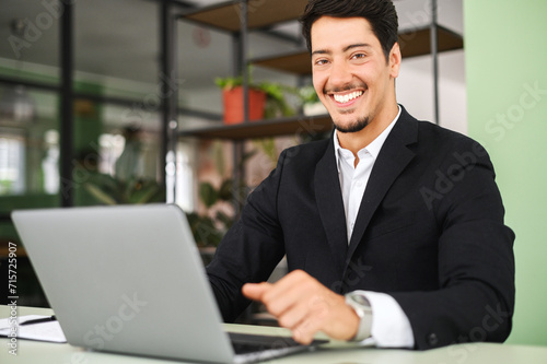 Cheerful clever male latin white collar worker using laptop indoors, looking at the camera and smiles, portrait of proud and happy male office employee on the workplace