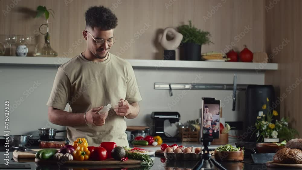 Young food blogger standing behind kitchen table, adding oil to dish and speaking in front on phone on tripod while filming cooking show for social media Young food blogger standing behind kitchen table, adding oil to dish and speaking in front on phone on tripod while filming cooking show for social media