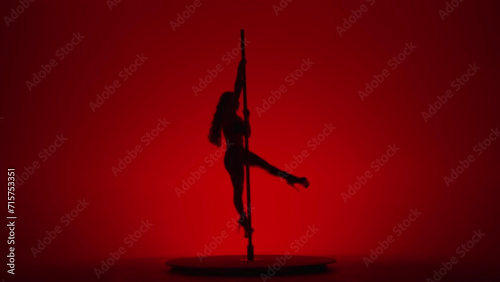 Dark silhouette of female dancer performing pole dance in studio on red background. Woman in high heeled shoes performs rotation on a pole.