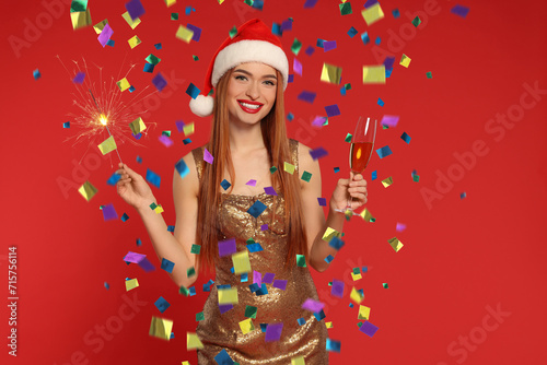 Happy woman in Santa hat with sparkler and glass of sparkling wine under falling confetti on red background