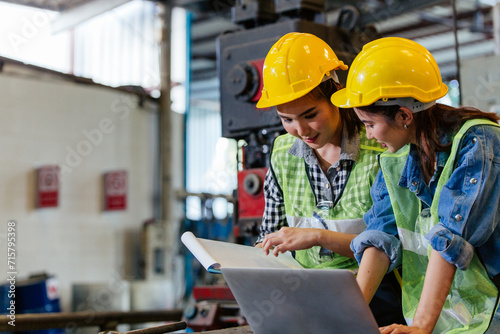Two female engineers or technician worker wearing safety hard helmet discuss project in industry manufacturing factory. woman using laptop checking work, team colleague note on checklist document photo
