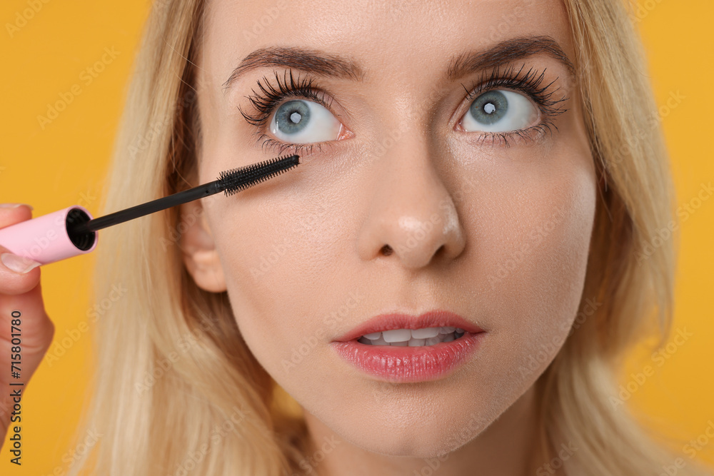 Beautiful woman applying mascara on orange background, closeup