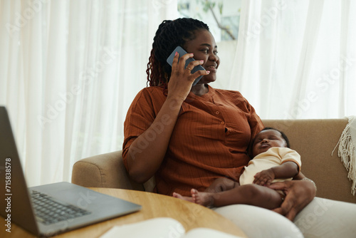 Positive Black woman with sleeping newborn baby talking on phone
