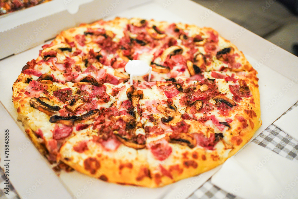 Freshly baked pizza with ham and mushrooms in an open box, cozy indoor setting.