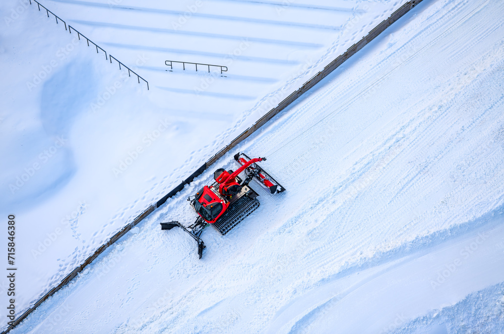 Red snow groomer in bright snowy landscape. Arial view of high tech vehicle preparing snow conditions for ski jumping in “Mühlenkopfschanze“ arena in Willingen, Germany. Seen from suspension bridge.
