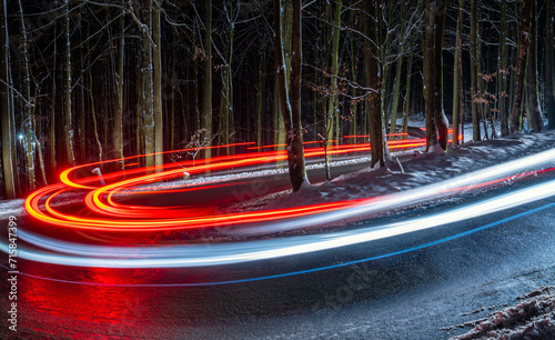 Country road at night with white and red light traces of cars driving through darkness. Long time exposure of a winding forest road with hairpin curve, reflecting wet street and snowy beech trees.
