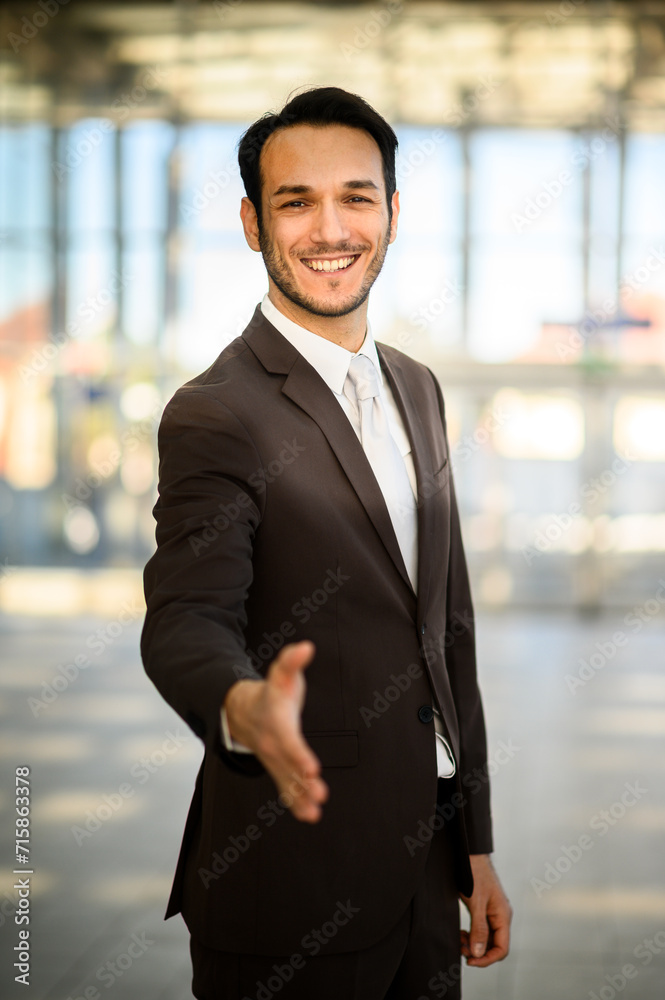 Smiling young man in suit extends a handshake, greeting at a bright modern office setting Smiling young man in suit extends a handshake, greeting at a bright modern office setting