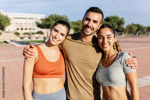 Portrait of three sportive friends smiling at camera embraced in an urban park in a sunny day