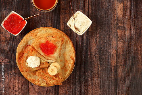 Delicious Russian pancakes with red caviar, soft cheese curd, bananas and honey on a wooden table. Healthy breakfast with ingredients, carnival concept and traditional national cuisine,