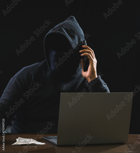 Hacker spy man oneperson black hoodie sitting on table hand holding mobile phone looking computer laptop used login password attack security data digital internet network system, night dark background photo