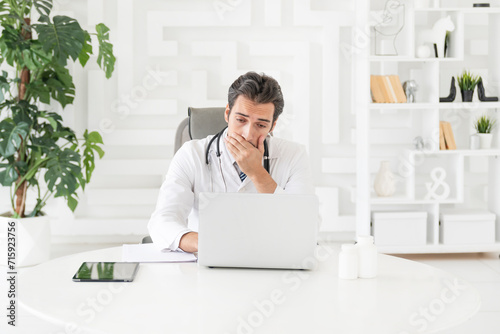Close up portrait of tired male doctor sitting at the desktop and working on laptop in the office of modern clinic 