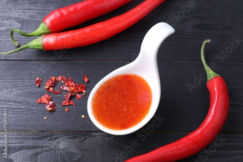 Spicy chili sauce in spoon and peppers on black wooden table