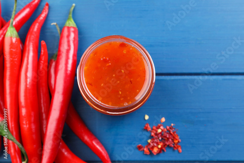 Spicy chili sauce in jar and peppers on blue wooden table, flat lay. Space for text