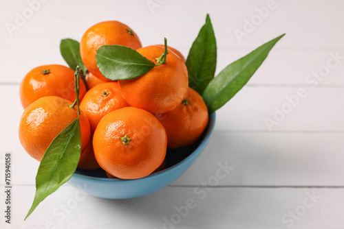 Fresh ripe juicy tangerines and green leaves in bowl on white wooden table, closeup. Space for text