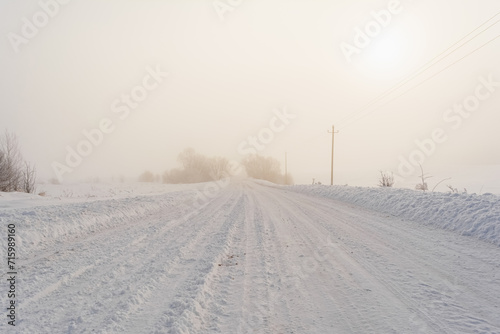 The dirt road has been cleared of snow. Poor visibility due to thick fog and snowstorm. Winter theme weather peculiarities. The sun in the haze on a frosty winter day