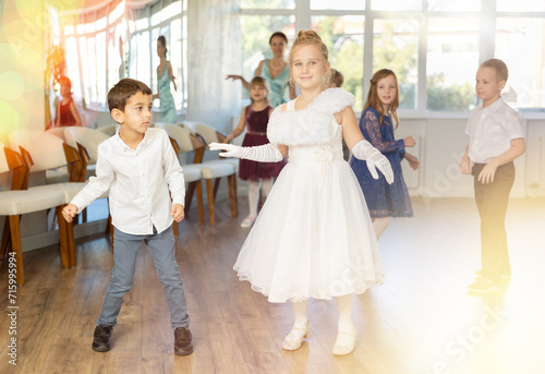 Smiling junior schoolchildren in party dresses learning dancing twist with pedagogue in social hall