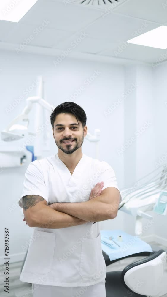 Vertical video of a smiling dentist standing arms crossed in modern clinic. Zoom in shot of young male dentist in medical uniform smiling and looking at camera while standing arms crossed in clinic Vertical video of a smiling dentist standing arms crossed in modern clinic. Zoom in shot of young male dentist in medical uniform smiling and looking at camera while standing arms crossed in clinic