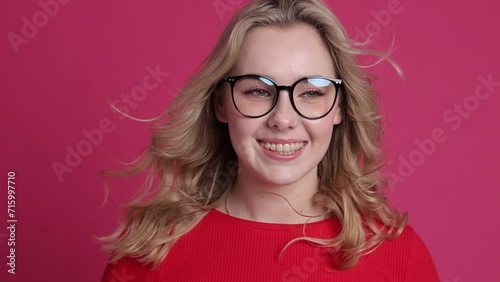Cheerful young caucasian woman with transparent ceramic braces on teeth isolated on pink backdrop. Blond girl in eyeglasses looks at camera. Windy effect with fan. Beauty, health and fashion concept. photo