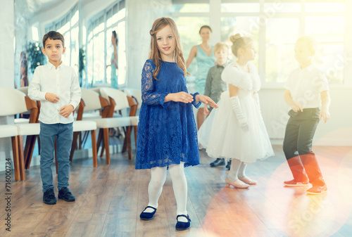 Cheerful preteen girl in blue lace dress dancing twist with classmates in bright classroom, full of energy and joy during school party ..