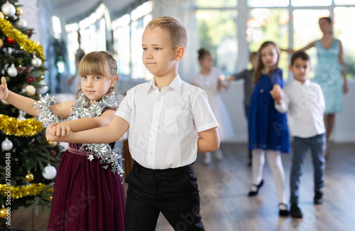 Couple of smiling tweens, boy and girl in festive clothing performing graceful waltz with group classmates under guidance of female teacher during Christmas event at school..
